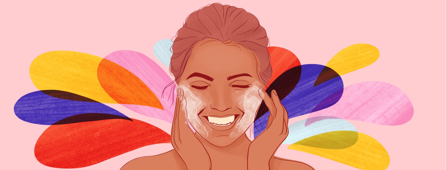 A woman rubs face wash into her cheeks, smiling, multicolored splashes in the background